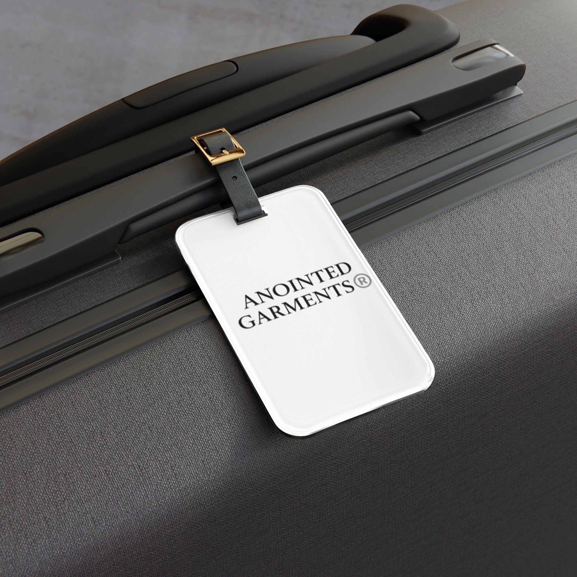 Anointed Garments white acrylic luggage tag with a black leather strap attached to a grey suitcase.