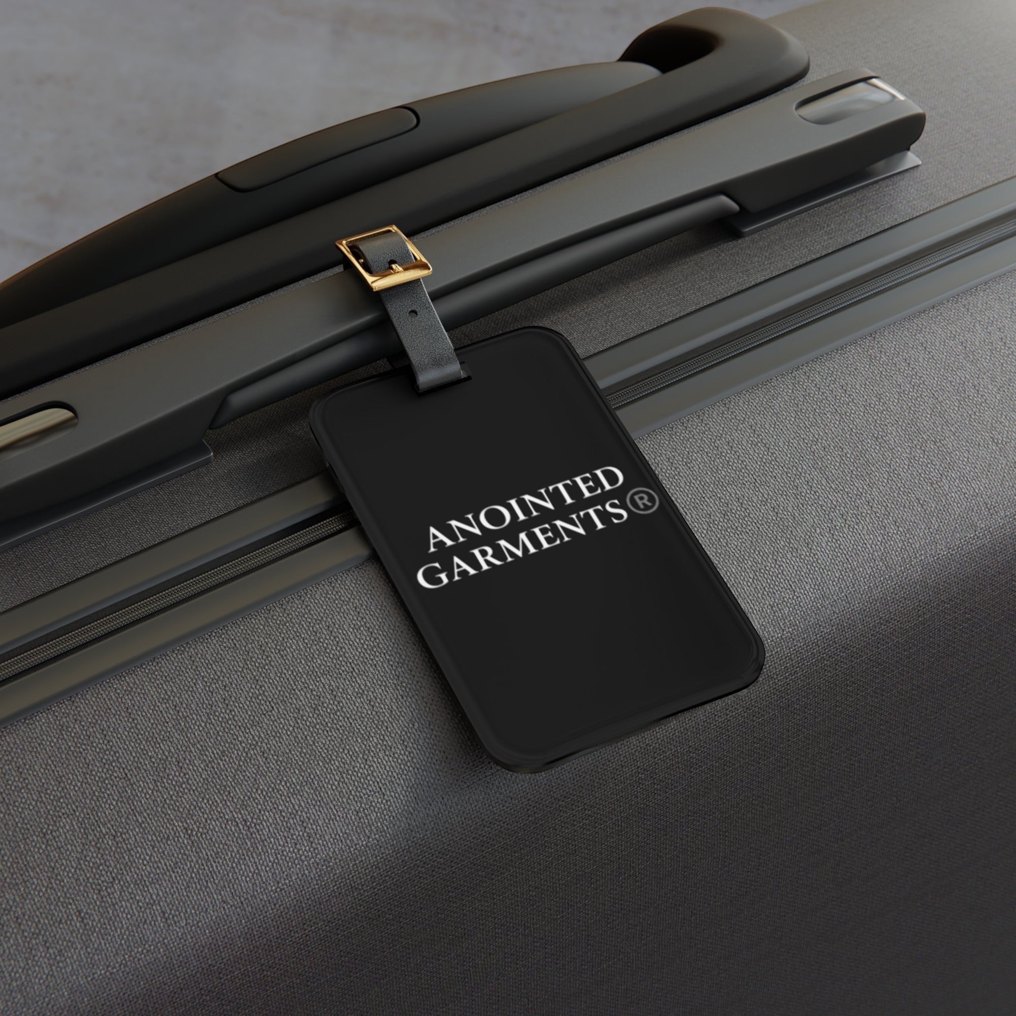 Anointed Garments black acrylic luggage tag with adjustable leather strap attached to a grey suitcase