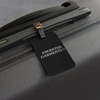 Anointed Garments black acrylic luggage tag with white logo and adjustable leather strap attached to a grey suitcase.