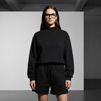 A woman wearing an Anointed Garments black mock neck pullover sweatshirt and matching shorts in a minimalist studio.