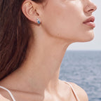 Anointed Garments blue clover moissanite stud earrings in platinum-plated sterling silver worn by a woman by the sea.