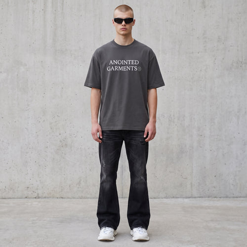 Model wearing an Anointed Garments charcoal grey boxy cotton t-shirt with white logo, black jeans, and sunglasses.