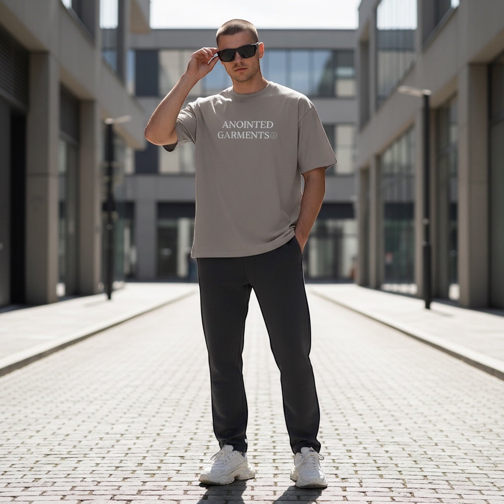 Model wearing Anointed Garments charcoal oversized cotton t-shirt with logo, sunglasses, and dark trousers outdoors.