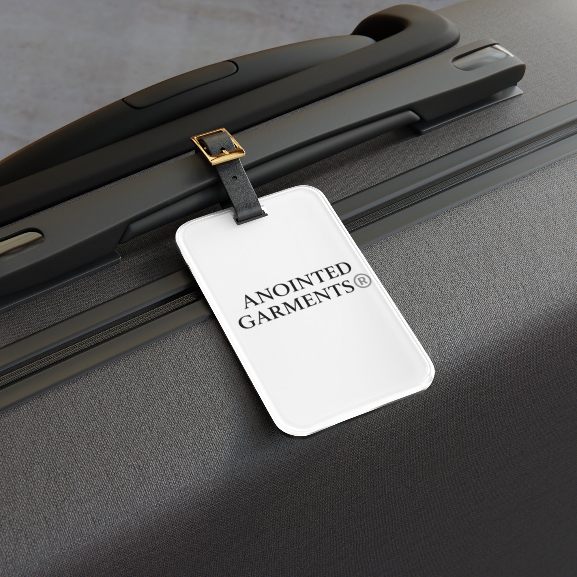 Anointed Garments white luggage tag with black logo attached to a textured black hardshell travel suitcase